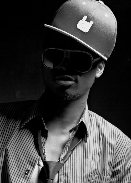 Monochrome close-up portrait of Josh wearing a flat brim cap and large sunglasses in deep studio shadow.