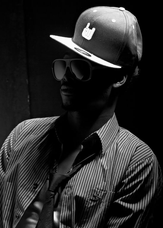Dramatic black and white studio portrait of Josh wearing a flat brim cap and large sunglasses in deep shadow.