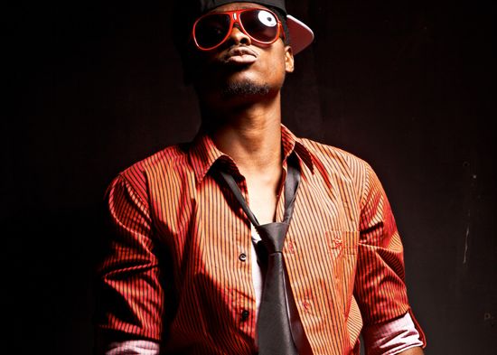 Urban studio portrait of Josh wearing a striped shirt, tie, and bold red-rimmed sunglasses against a dark background.