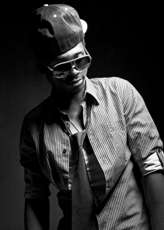 Monochrome studio portrait of Josh looking downward while wearing a backward cap, striped shirt, and tie.